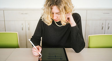 A woman sits at a table in front of her tablet and writes something on the tablet with a pen.