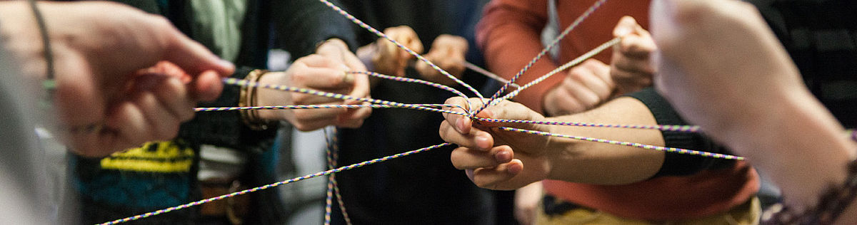 Several people stand together in a circle holding pieces of string that overlap in the centre of the circle.